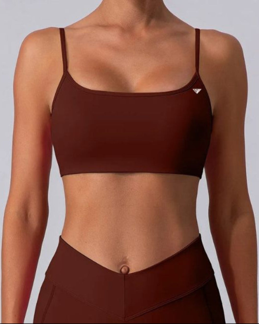 A woman wearing a dark brown scoop-neck sports bra and matching high-waisted leggings against a light gray studio background.
