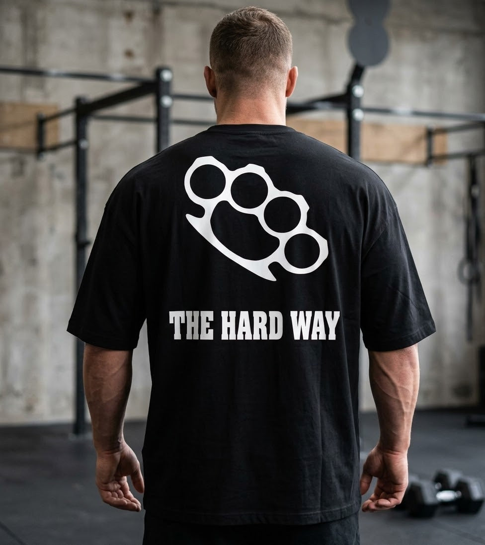 A man viewed from behind wearing a black t-shirt with a white brass knuckles graphic and the text 'THE HARD WAY' printed on the back, set in a gym environment.